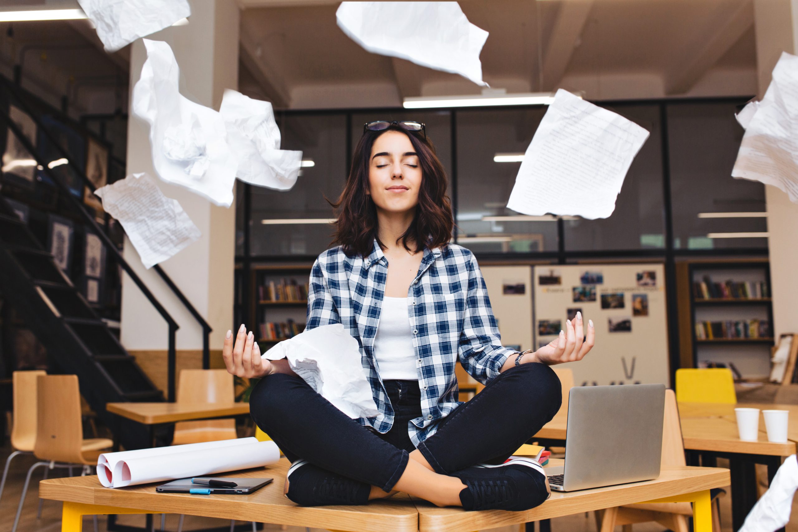 Young pretty joyful brunette woman meditating on table surround work stuff and flying papers. Cheerful mood, taking a break, working, studying, relaxation, true emotions.
